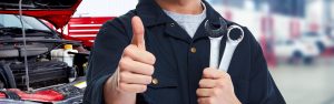A mechanic giving a thumbs up while holding two wrenches with a car's engine in the background.