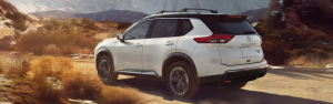 A white Nissan Rogue SUV driving on an off-road path with mountains in the background.