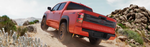 Red pickup truck driving uphill on a rocky off-road trail in a desert landscape.
