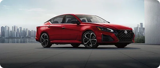 2026 Nissan Altima SV in red parked on a waterfront plaza with a city skyline behind it, front three-quarter view