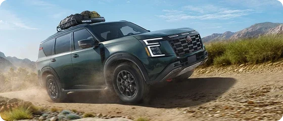 2026 Nissan Armada PRO-4X 4WD in green driving on a dusty desert trail with a loaded roof rack, front three-quarter view
