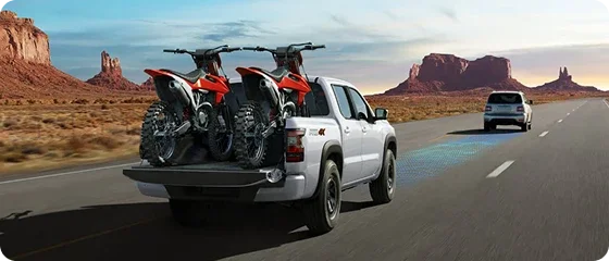 2026 Nissan Frontier PRO-4X 4x4 in white carrying two dirt bikes in the bed on a desert highway, rear three-quarter view