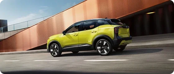 2026 Nissan Kicks SV AWD in yellow driving past a corrugated orange building, rear three-quarter view