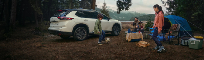 White Nissan SUV parked at a forest campsite while a family plays soccer