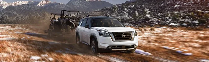 White 2026 Nissan Pathfinder towing a side-by-side on a trailer across open country