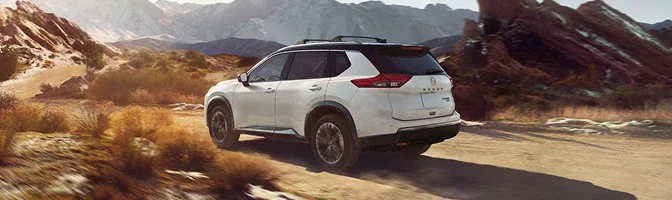 White 2026 Nissan Rogue with a roof rack driving a desert road below mountains
